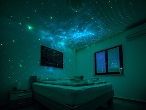 Un dormitorio con cama, mesita de noche, televisor en la pared y ventana.