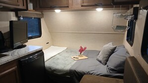 Interior de una autocaravana compacta, con kitchenette, cama con almohadas, una mesita con un florero y un televisor montado.