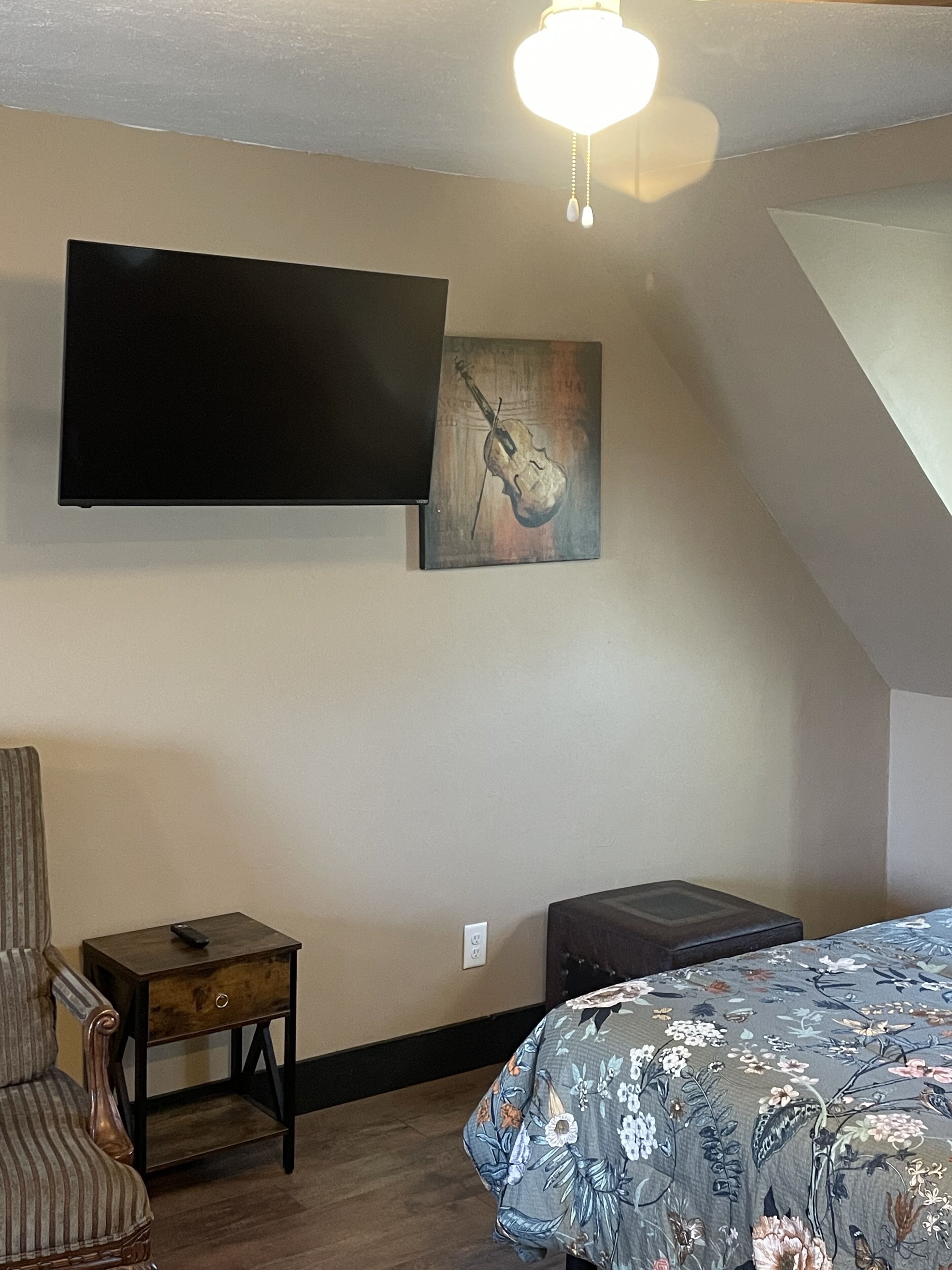 Een slaapkamer met een bed, een aan de muur bevestigde televisie, een gitaar schilderij, een stoel en een klein tafeltje.