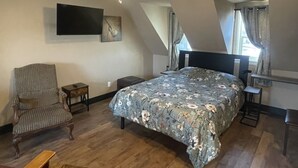Un dormitorio con cama, una silla, una mesita pequeña, un ventilador de techo y un televisor montado en la pared.