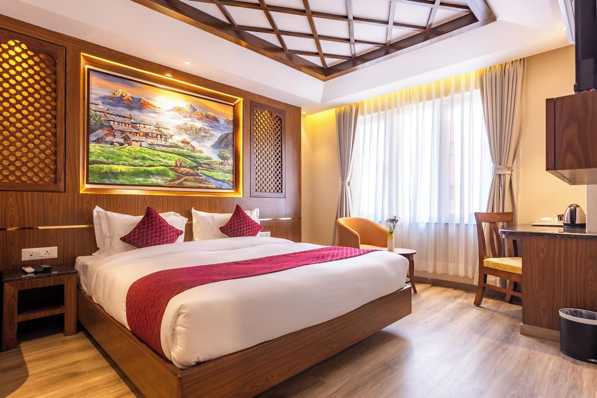 Ein Hotelzimmer mit einem großen Bett, einem Holzkopfstück, einem Landschaftsbild, einem Schreibtisch mit Stuhl und einem Fenster mit Vorhängen.