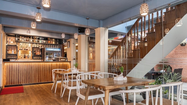 Un restaurante con zona de bar, mesas de madera, sillas blancas y una escalera.