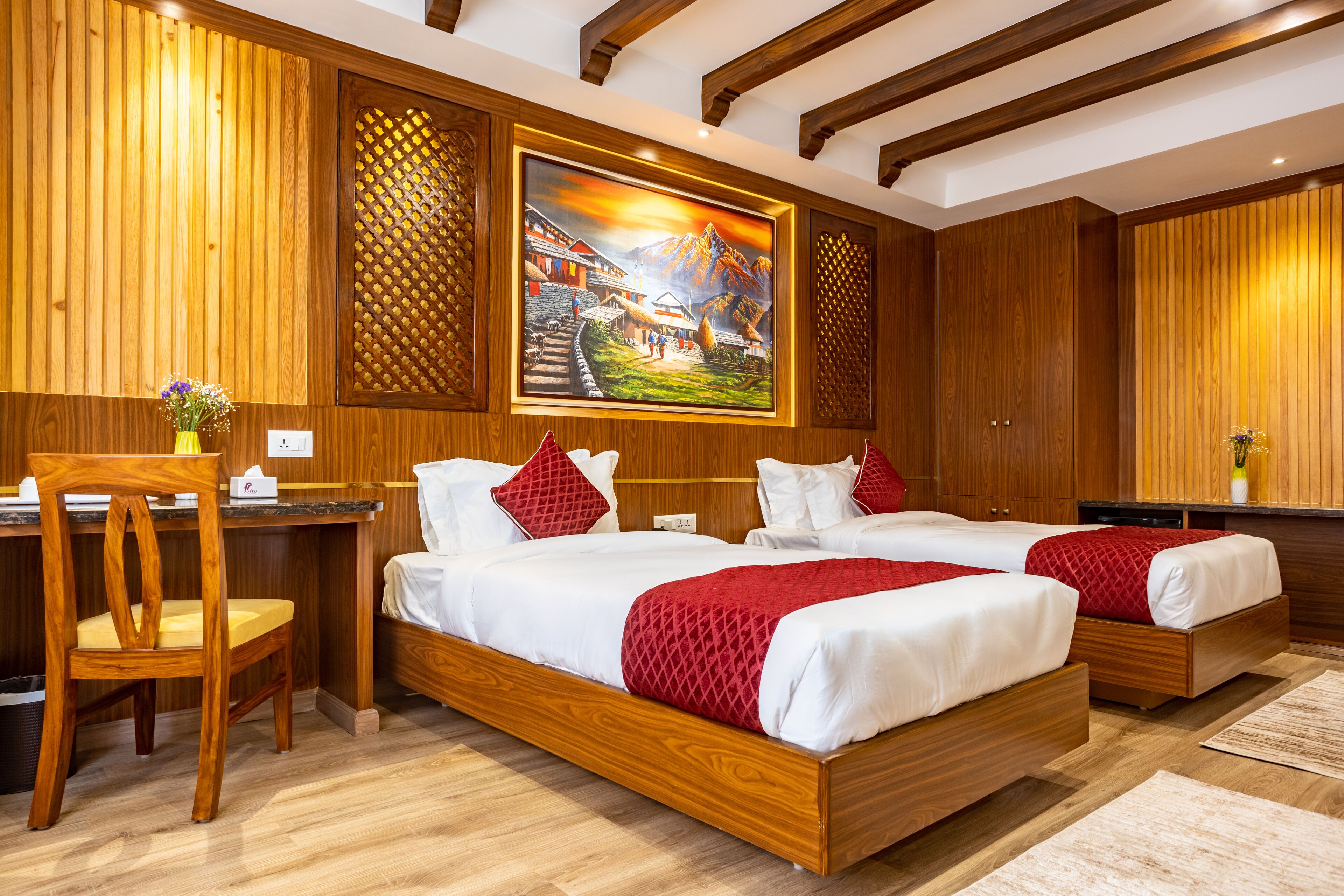 Ein Hotelzimmer mit Holzvertäfelung, einem Schreibtisch, zwei Betten mit rot-weißer Bettwäsche und einem Gemälde einer Berglandschaft an der Wand.