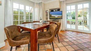 A dining area with a wooden table and cushioned chairs, a flat-screen TV mounted above it, and large windows offering a view of the outdoors. - Senmare - Geschmackvolles Ferienhaus für Sechs Personen in Wattnähe!! (Sylt)