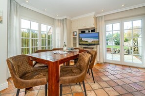 Ein Essbereich mit Holztisch und gepolsterten Stühlen, einem flachbildfernseher darüber und großen Fenstern mit Blick ins Freie.