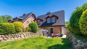 A two-story house with a stone retaining wall, a patio with outdoor furniture, and a well-maintained garden. - Senmare - Geschmackvolles Ferienhaus für Sechs Personen in Wattnähe!! (Sylt)