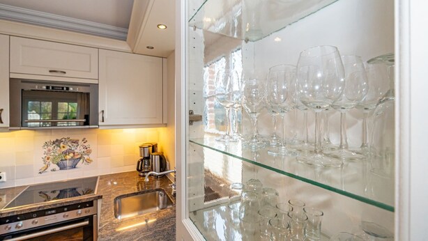 A kitchen with a built-in oven, a coffee maker, and a glass display case with wine glasses. - Senmare - Geschmackvolles Ferienhaus für Sechs Personen in Wattnähe!! (Sylt)