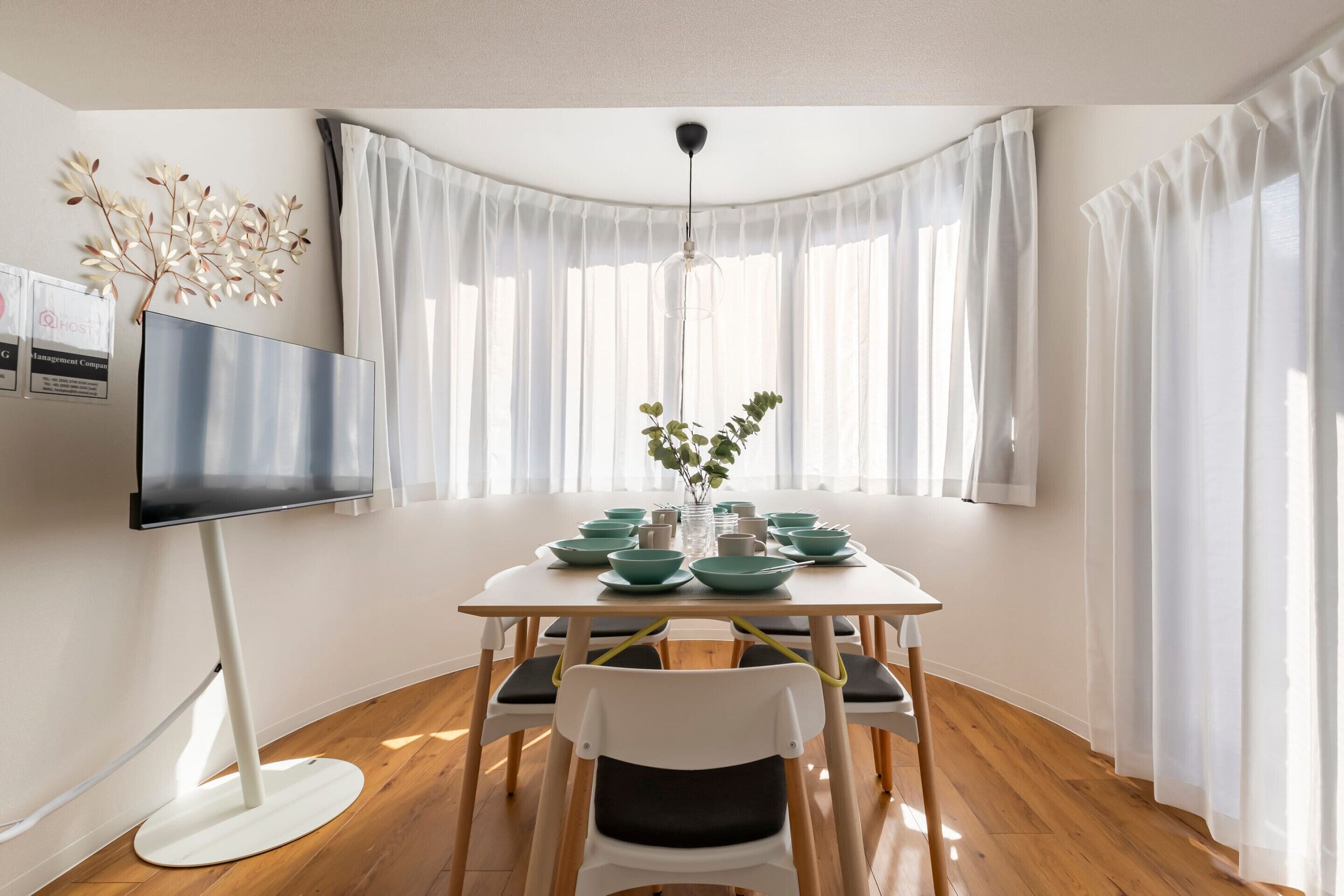 A dining area with a wooden table, white chairs, a flat-screen TV, and a vase with green leaves.