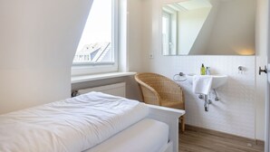 Ein kleiner, heller Raum mit einem Einzelbett, einem Korbsessel, einem weißen Waschbecken und einem großen Spiegel.