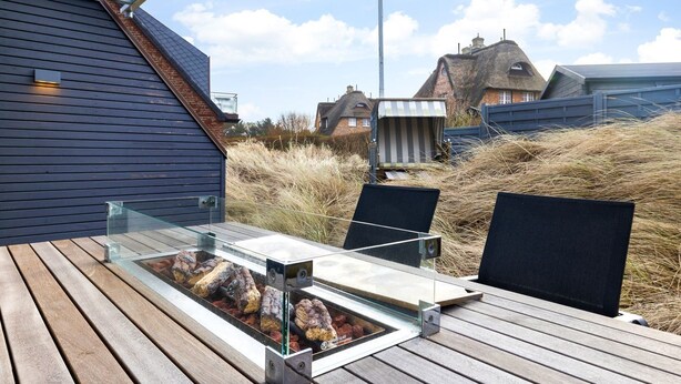 Een eethoek buiten met een tafel met glazen blad, metalen stoelen en een barbecue met eten erop.