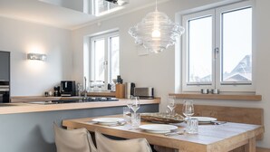 Een moderne keuken met een eethoek, met een houten tafel gedekt voor twee personen, een hanglamp en uitzicht buiten door grote ramen.