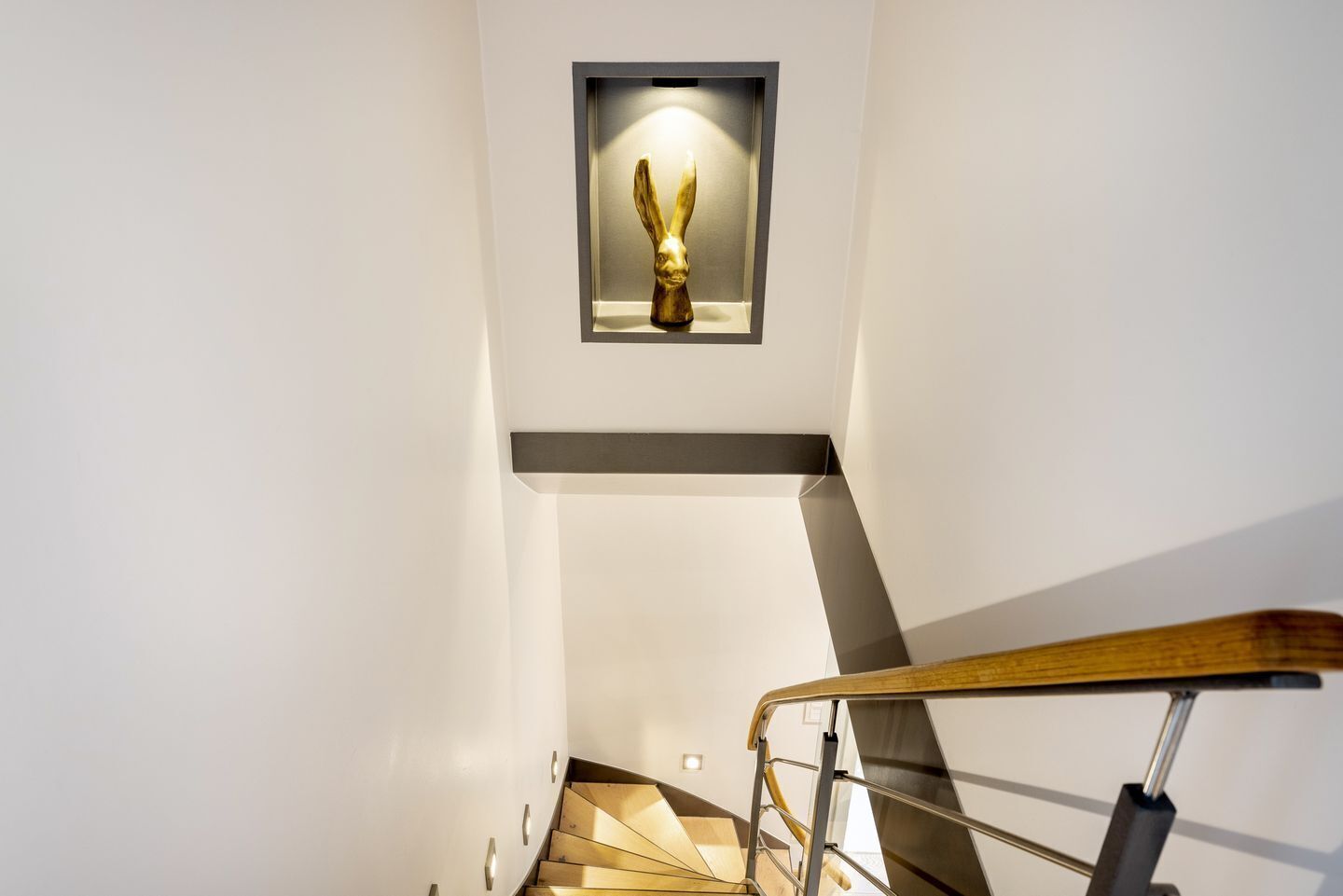 A modern staircase with a wooden handrail and a golden sculpture in a recessed alcove.