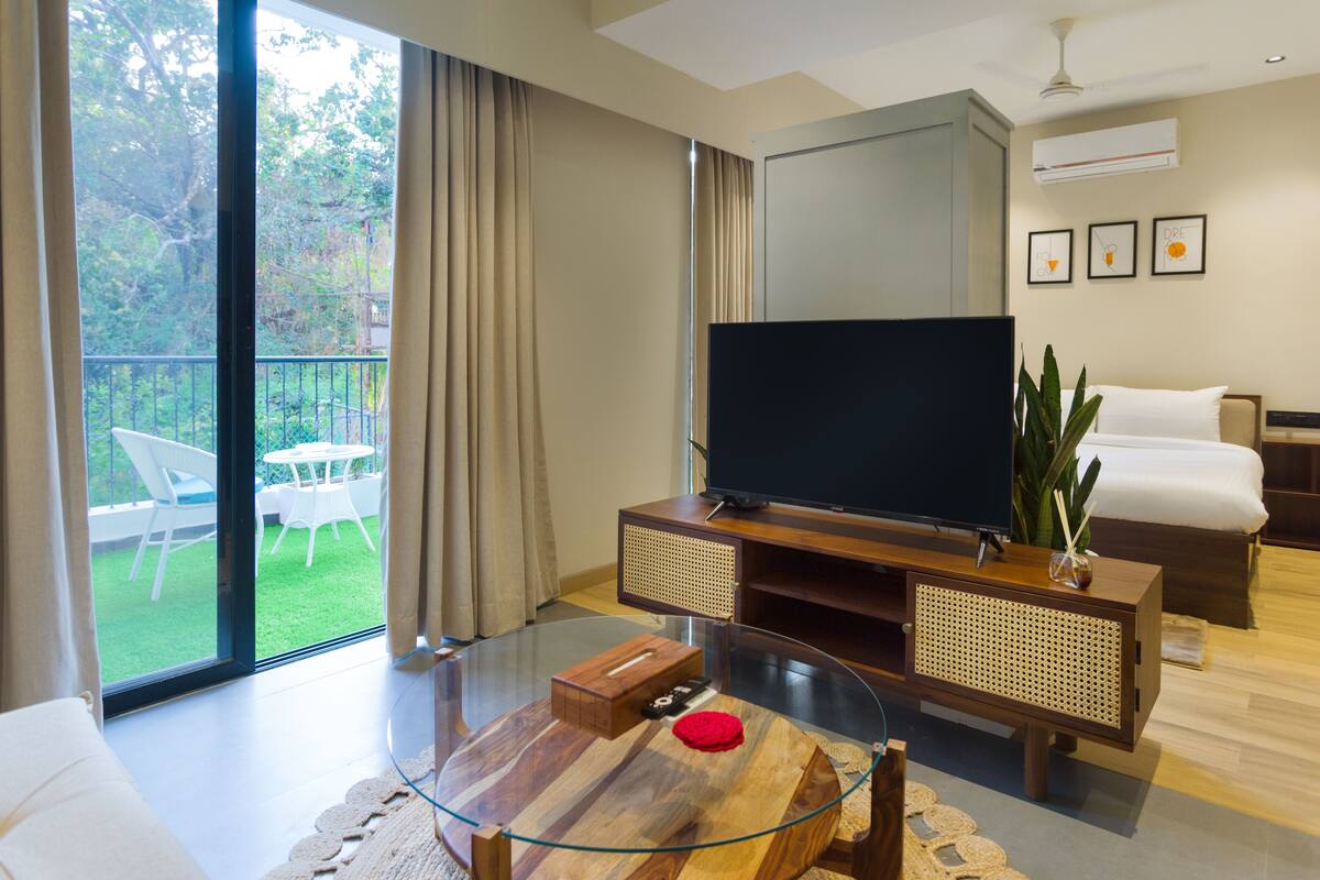 A modern living room with a glass coffee table, a flat-screen TV, a sofa, and a balcony with outdoor furniture.