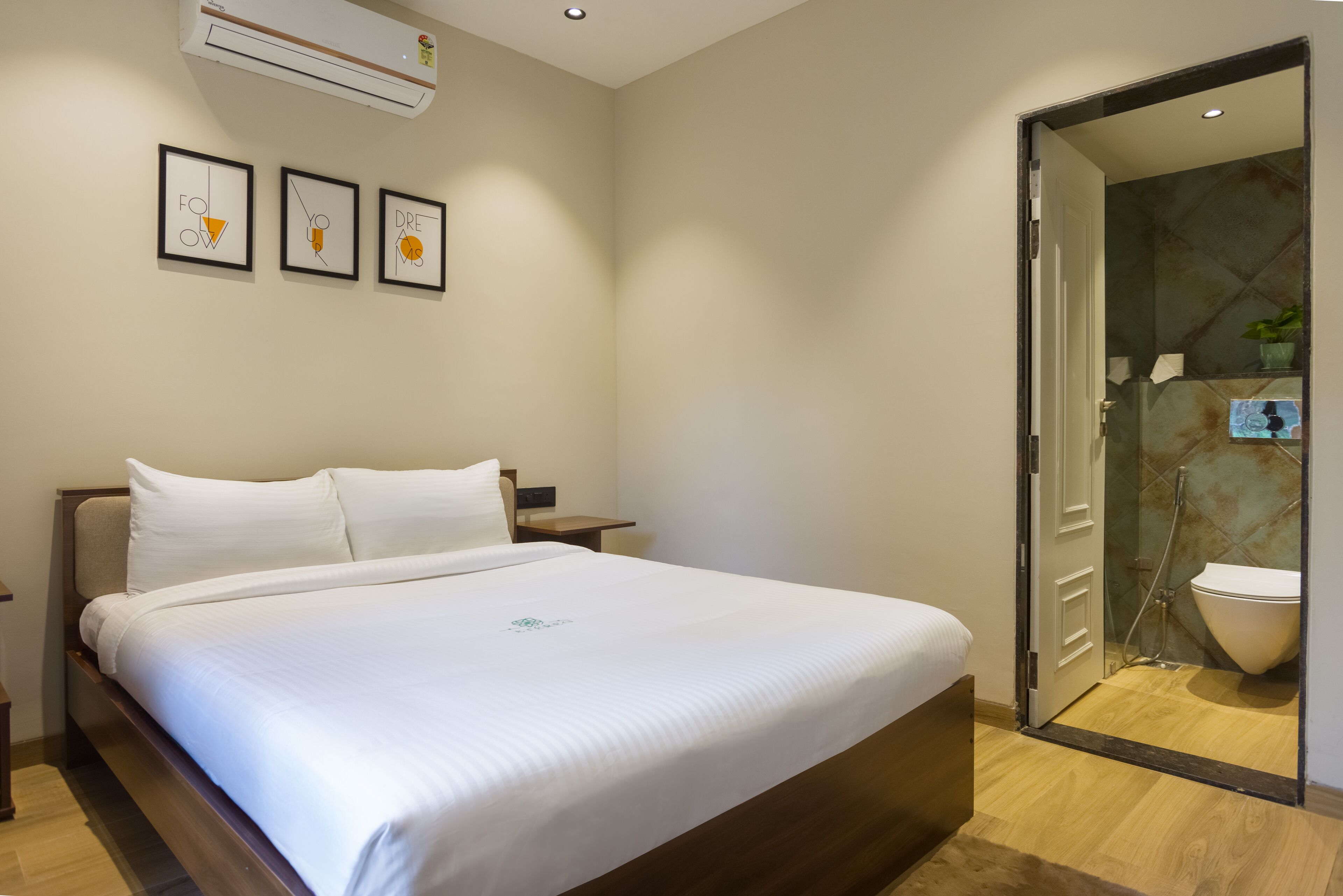 A hotel room with a large bed, a bedside table, and a wall-mounted air conditioner. There is a door leading to a bathroom with a shower and toilet.