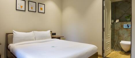 A hotel room with a large bed, a bedside table, and a wall-mounted air conditioner. There is a door leading to a bathroom with a shower and toilet.