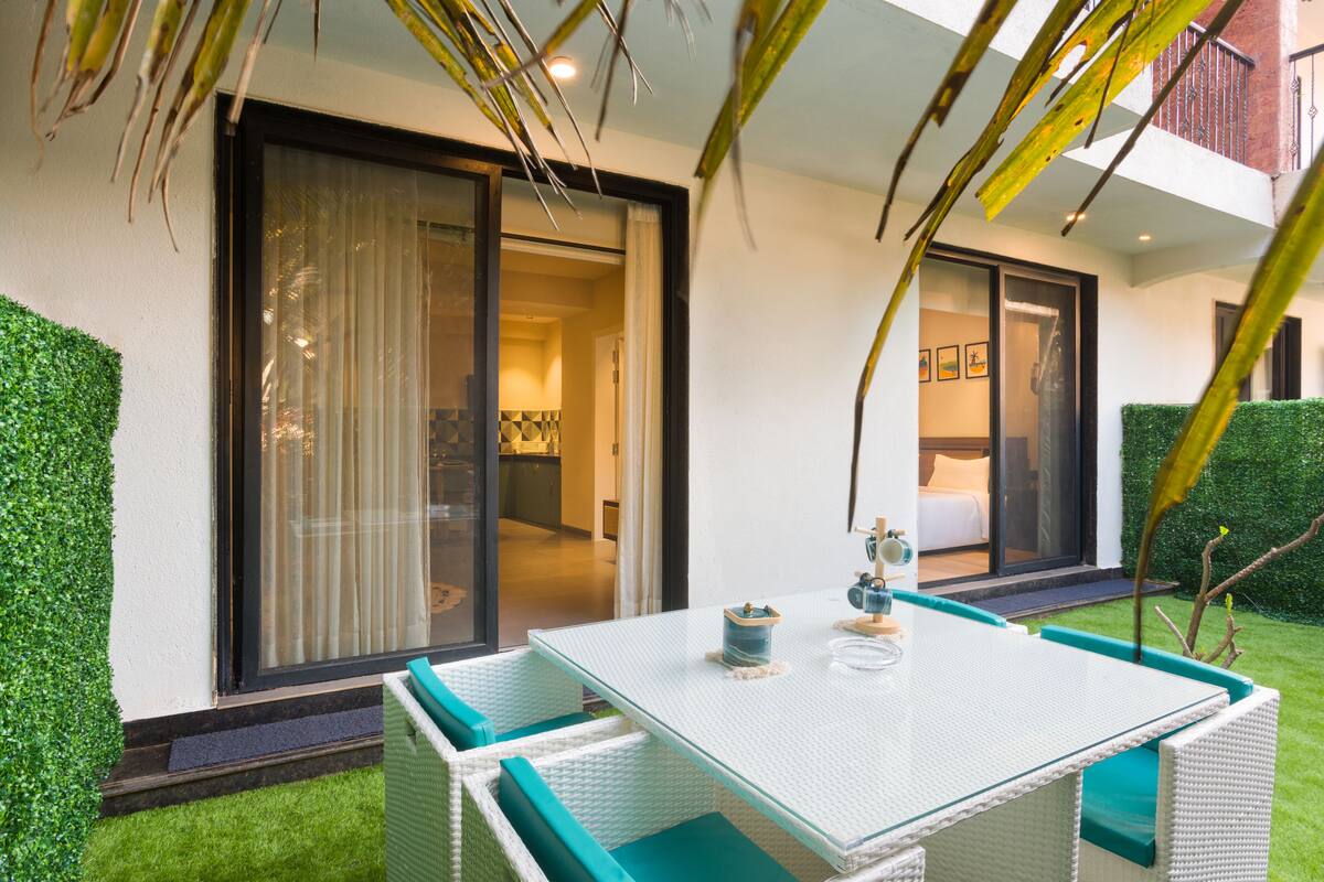 A modern outdoor seating area with a glass table and wicker chairs, a sliding glass door leading to a bedroom, and a well-maintained lawn.