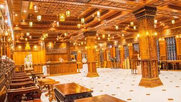 A spacious hall with ornate wooden columns and intricate ceiling designs, featuring rows of seating and tables.