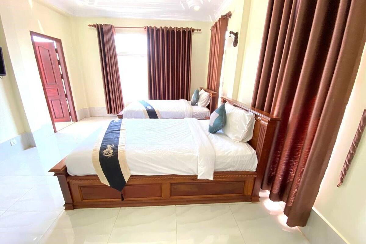 Une chambre d’hôtel avec deux lits, une tête de lit en bois, une grande fenêtre avec des rideaux et un sol carrelé.