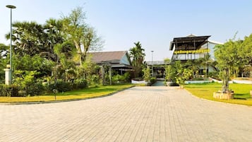 A paved pathway leading to a building with a traditional roof, surrounded by lush greenery and palm trees.