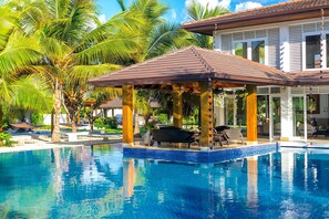 A swimming pool with a covered seating area, palm trees, and a building with large windows. - Srvittinivillas Agp36 / Spacius / Confortable Luxury Villa/ Family/ Team/ Cdc (La Romana)