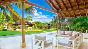 A covered outdoor seating area with white furniture and a view of a lush garden and a house in the background.