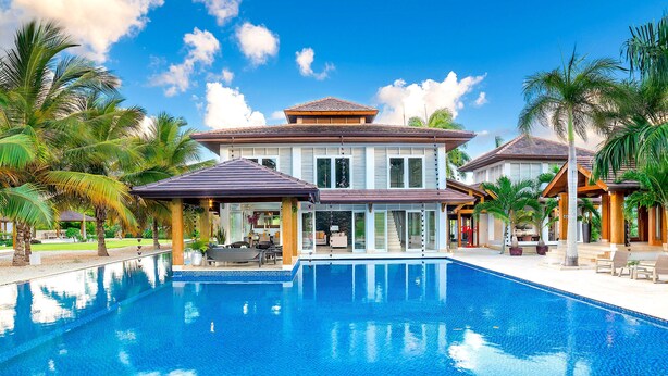 A large swimming pool with a house in the background, surrounded by palm trees and clear skies.
