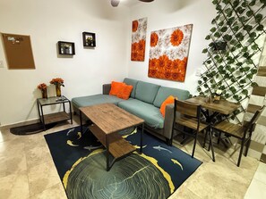 A living room with a sofa, coffee table, and a dining area with chairs. There are wall-mounted pictures and a plant on a trellis. - Playas y Arenas (Puerto Plata)