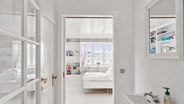 Een lichte, witte kamer met een bed, een nachtkastje en een raam. Er is een deur die leidt naar een andere kamer met een boekenkast en een raam.