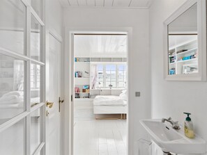 A bright, white room with a bed, a bedside table, and a window. There is a door leading to another room with a bookshelf and a window.