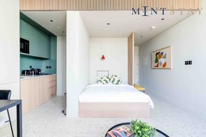 A modern hotel room with a kitchenette, a bed with a wooden headboard, a small table, and a potted plant.