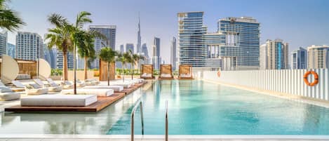 A rooftop infinity pool with lounge chairs and palm trees, overlooking a city skyline with modern buildings.