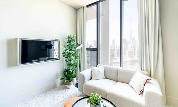 Un salon moderne avec un canapé, un téléviseur à écran plat, une plante en pot et une grande fenêtre avec des rideaux.
