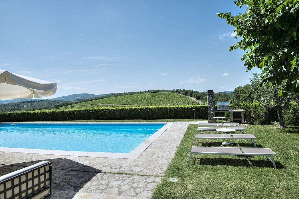 Une piscine dotée de chaises longues et d’un espace barbecue, le tout entouré de haies bien entretenues et offrant une vue sur des collines ondulantes.