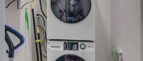 Un lavadero con dos lavadoras apiladas de carga frontal, un trapeador, un cubo y un radiador.
