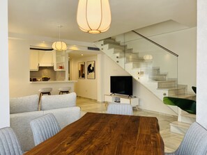 Un salon moderne avec une table à manger en bois, des canapés gris, un téléviseur et un escalier doté de rampes en verre.