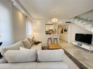Una sala de estar moderna con sofá, una mesa de centro y televisión. Cuenta con una zona de cocina con barra y taburetes.