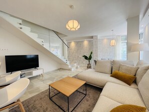A modern living room with a flat-screen TV, a wooden coffee table, a sofa with cushions, and a staircase with glass railings. - 3010 Spacious Luxury Family Apartment in Marbella's Heart (Marbella)