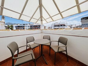 Un espace extérieur abrité, avec une table et des chaises, offrant une vue sur un quartier résidentiel, avec des bâtiments et des montagnes en arrière-plan.
