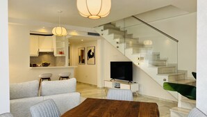 A modern living room with a wooden dining table, grey sofas, a TV, and a staircase with glass railings. - 3010 Spacious Luxury Family Apartment in Marbella's Heart (Marbella)