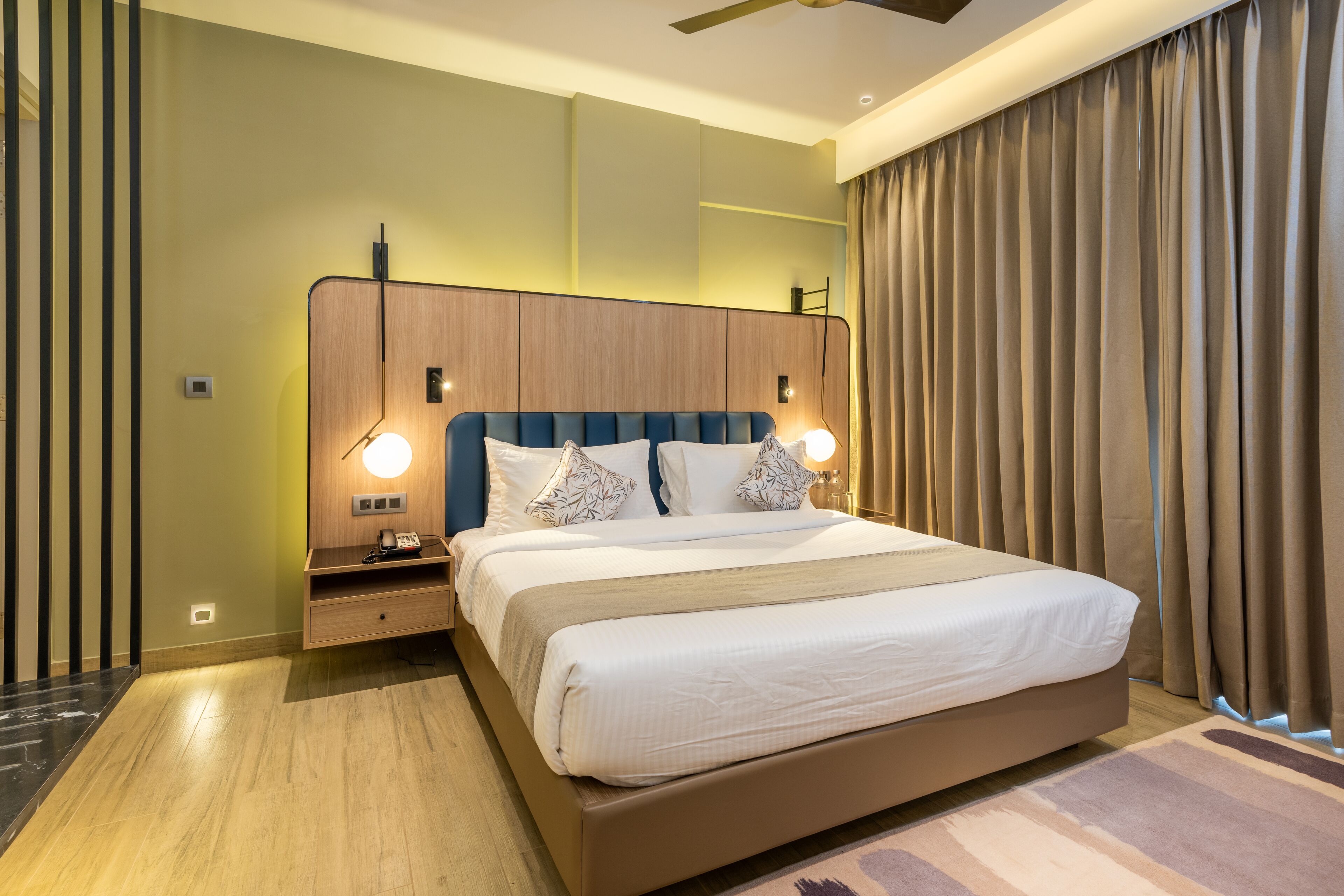 A modern hotel room with a large bed, wooden headboard, bedside lamps, and a view of the city through large windows.