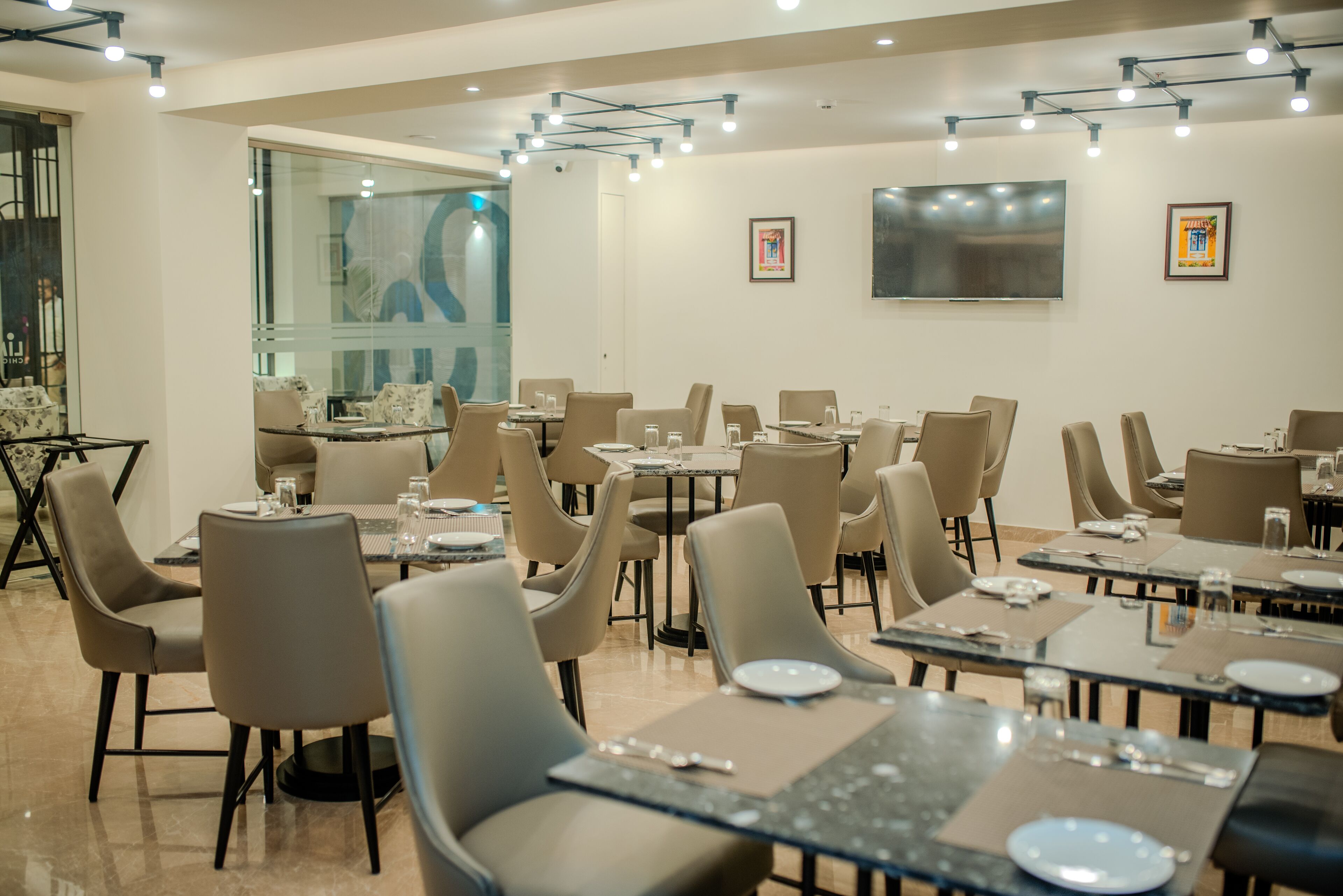 Un espace repas moderne avec plusieurs tables et chaises, un grand téléviseur fixé au mur et une cloison en verre.