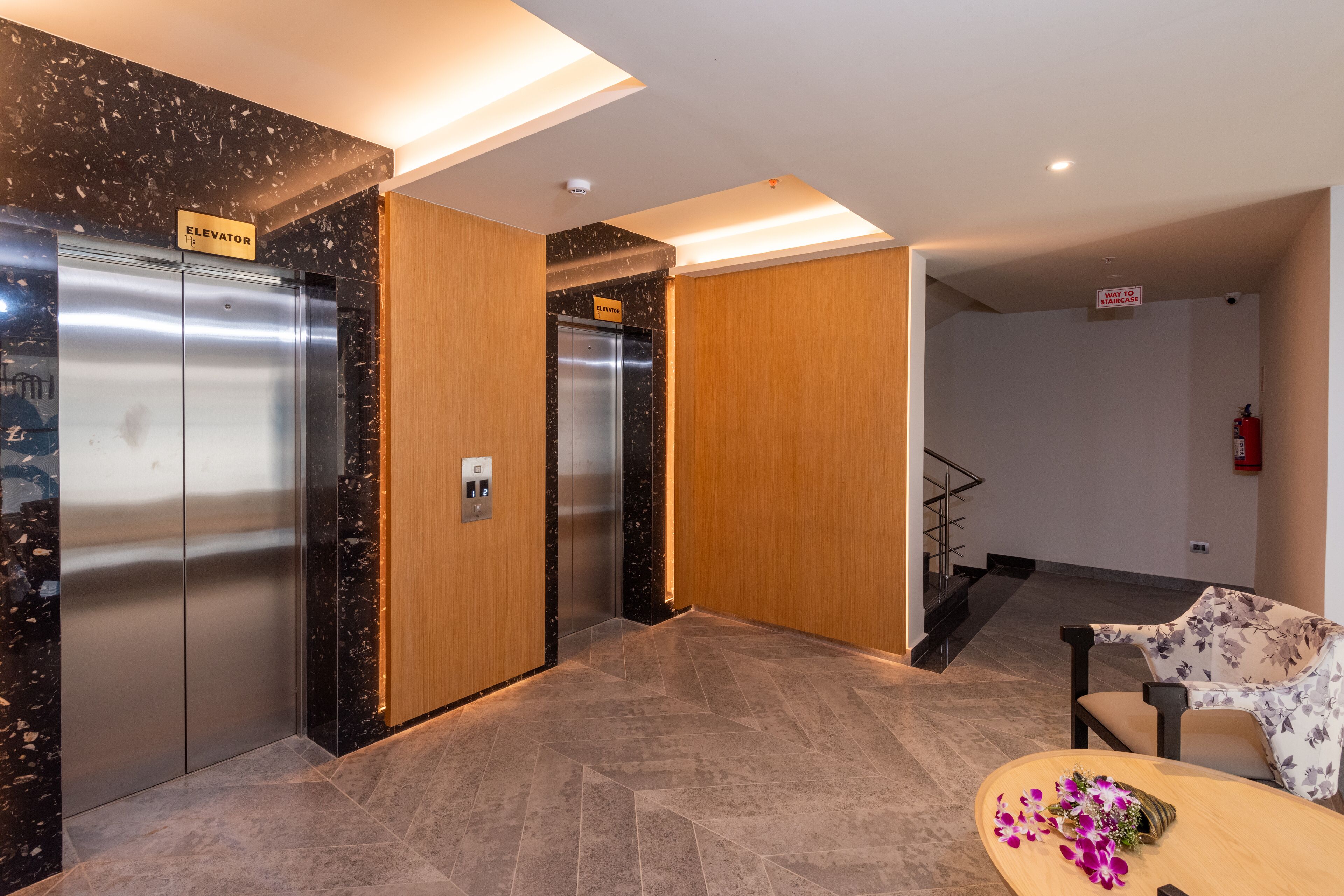 A modern lobby with two elevators, a wooden reception desk, and a staircase.
