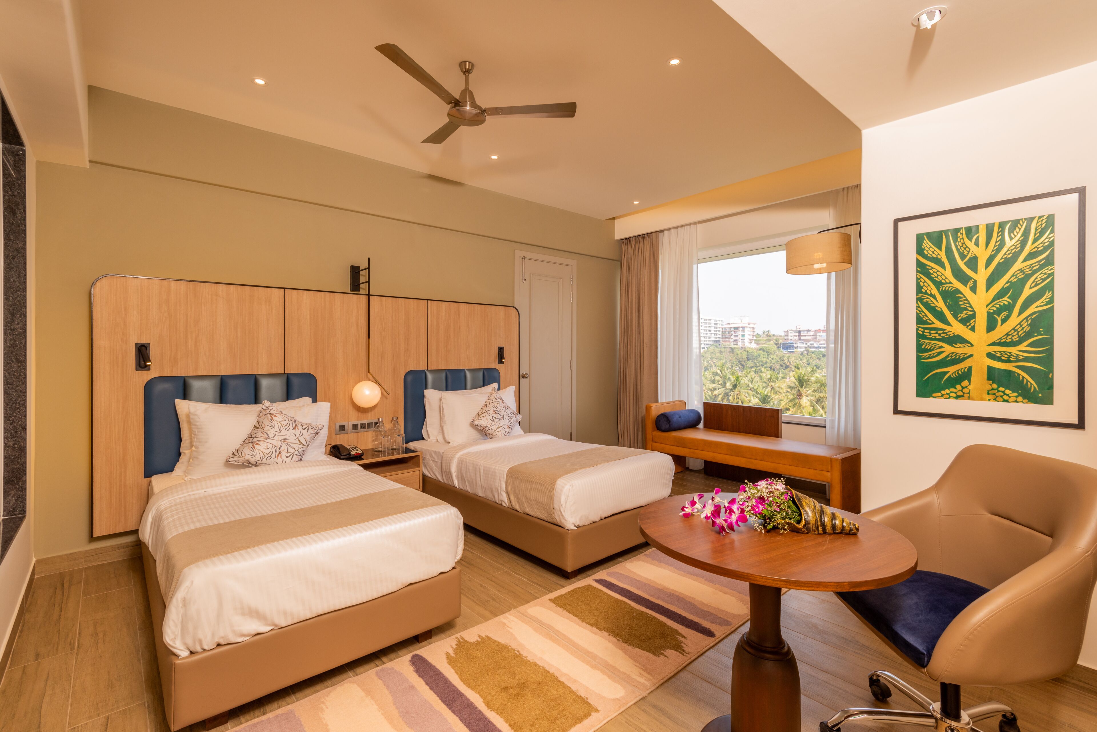 Habitación de hotel con dos camas, un escritorio, una silla, un ventilador de techo y un ventanal con vista a la ciudad.