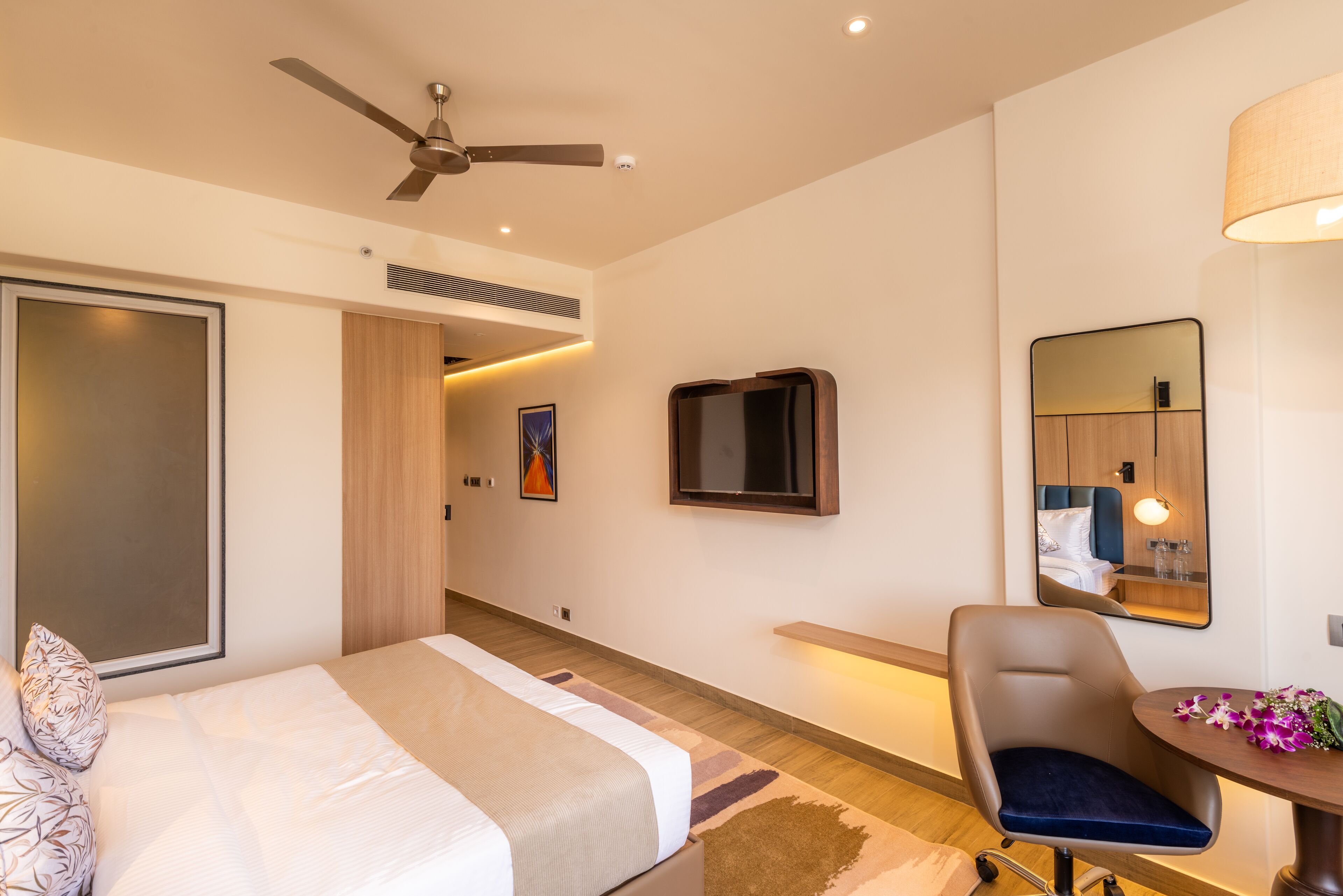 Una habitación de hotel moderna con cama, escritorio, silla, ventilador de techo, televisor y espejo.