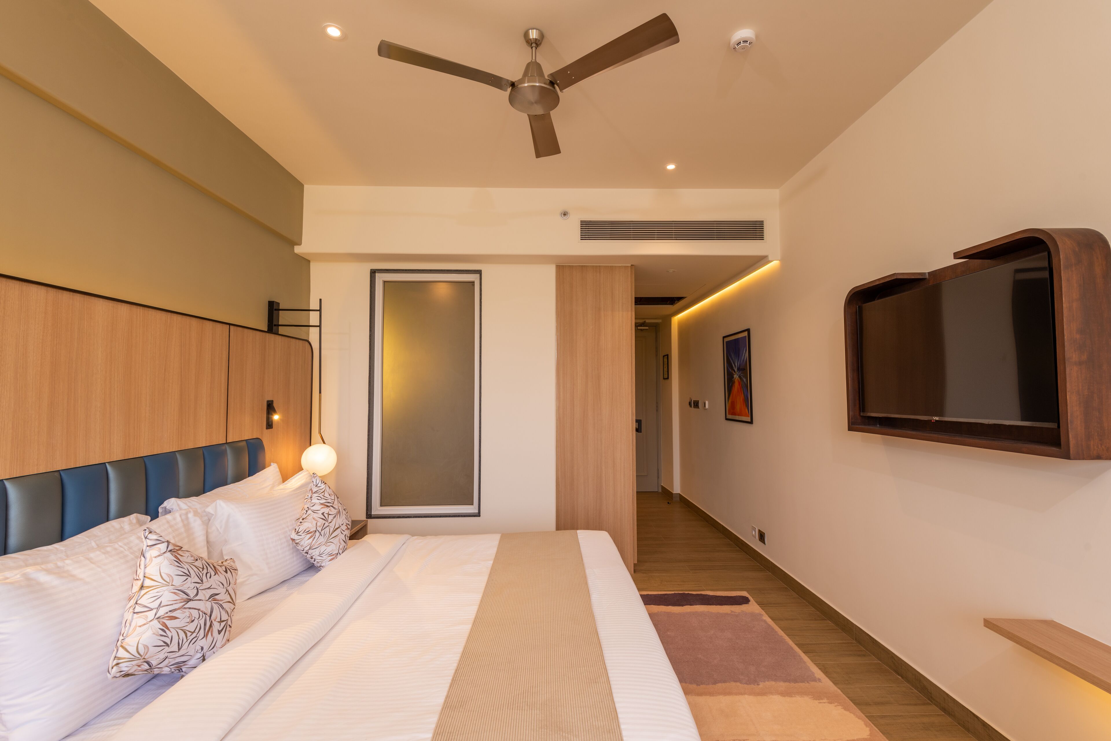 A modern bedroom with a bed, a ceiling fan, a TV mounted on the wall, and a wooden wardrobe.
