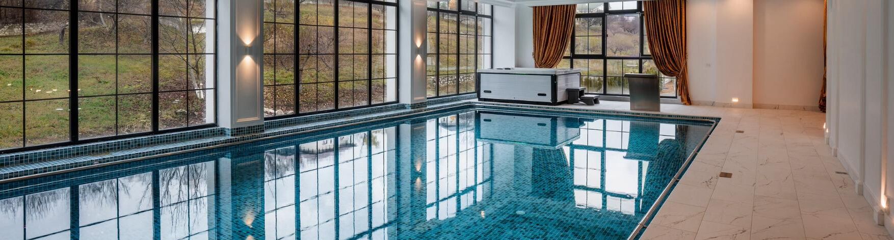 A large indoor swimming pool with clear blue water, surrounded by large windows offering views of a grassy area.