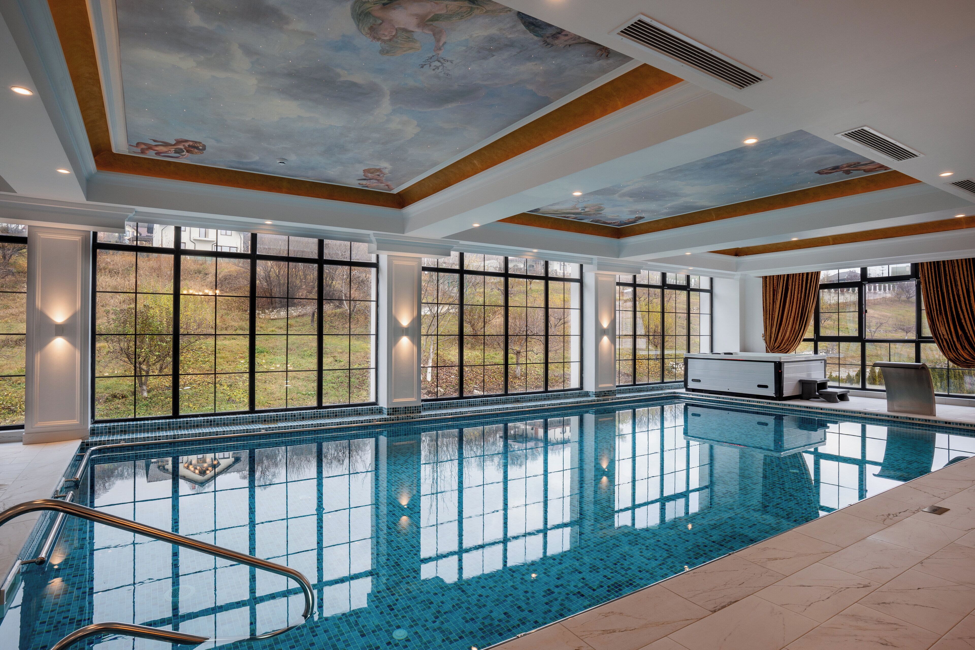 A large indoor swimming pool with clear blue water, surrounded by large windows offering views of a green landscape.