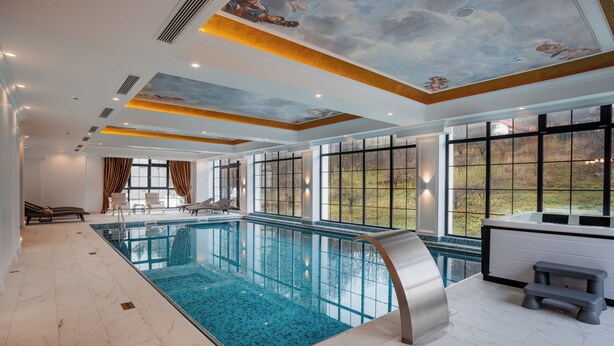 Una gran piscina interior con agua azul cristalina, un mural en el techo y grandes ventanales que ofrecen vistas al exterior.