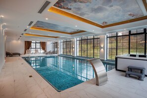 Una gran piscina interior con agua azul cristalina, un mural en el techo y grandes ventanales que ofrecen vistas al exterior.