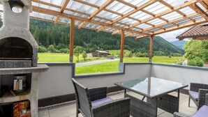 A covered patio area with a stone-built grill, wicker furniture, and a view of a green landscape with mountains. - Apartment Neukirchen near Ski Slopes (Neukirchen am Großvenediger)